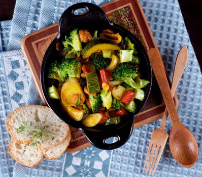 Homemade Colorful Vegetables Ragout with Zucchini, Carrots, Broccoli, Leek and Red Bell Pepper in Black Iron Stewpot with Wooden Spoon and Fork and Bread closeup on Blue Napkin. Top View