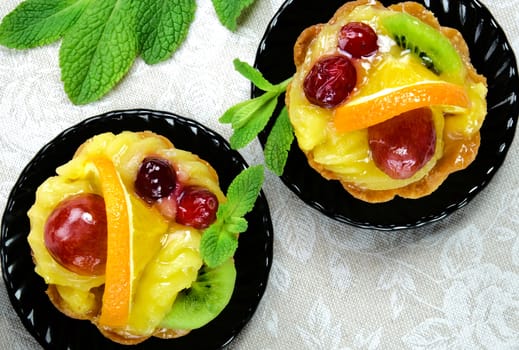Cakes with slices of fresh fruit orange, kiwi and grapes with jelly and cream on a black saucers on a table cloth.