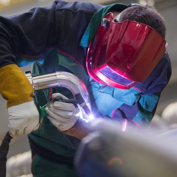 Industrial worker with protective mask welding inox elements in steel structures manufacture workshop.