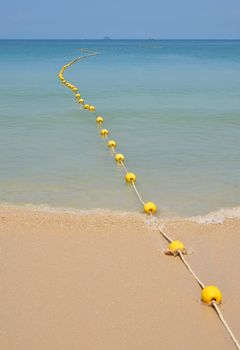 Chain of yellow polystyrene sea marker buoys with cable tow at sand beach and in blue sea water with clear sky above, perspective