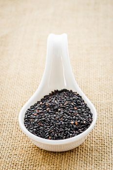 Black sesame seeds in white spoon on sack background.