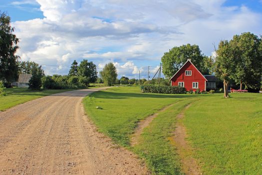 A quiet village road lined with farmhouse. Green trees and blue, cloudy sky in background.