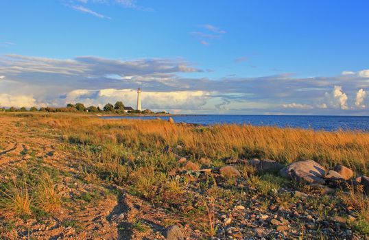 Stony shore with dry grass. Old lighthouse, blue sky and clouds in background. Evening sunlight in autumn.  Kihnu island, Estonia.