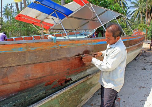 A fisherman works on fixing his longtail boat by caulking the joints of the hull of his boat with traditional materials and techniques.Thong Krut, Koh Samui, Thailand .