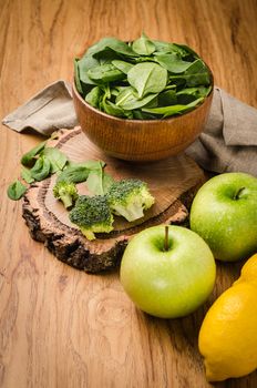 Spring spinach leaves in the bowl, broccoli, lemons and apples on wooden table background