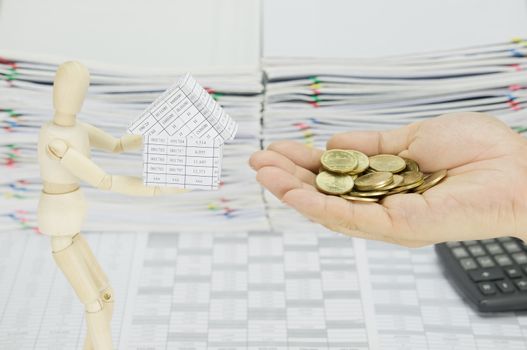 Man holding gold coins to buy house from wooden dummy on finance account have blur calculator and overload of paperwork as background.