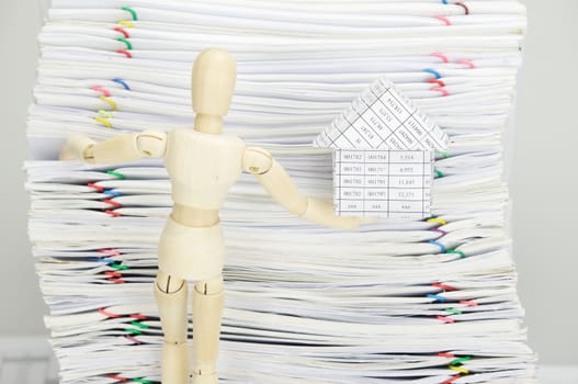 Wooden dummy holding house and climb on overload of paperwork with colorful paperclip as background.