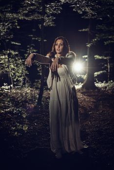 Young woman walking through the forest at night in white dress with outstretched arms in front of body.
