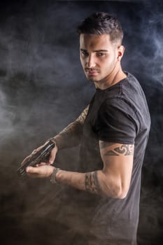 Young handsome man holding a hand gun, wearing black t-shirt, on dark background in studio