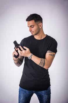 Young handsome man holding a hand gun, wearing black t-shirt, on light background in studio