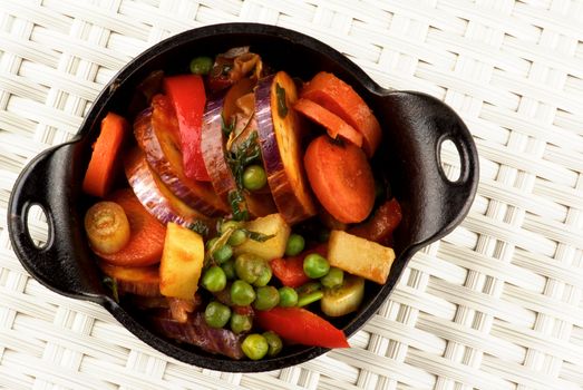 Delicious Homemade Colorful Vegetables Ragout with Eggplant, Carrots, Potatoes, Leek, Red Bell Pepper and Green Pea in Black Iron Stewpot closeup on Wicker background. Top View