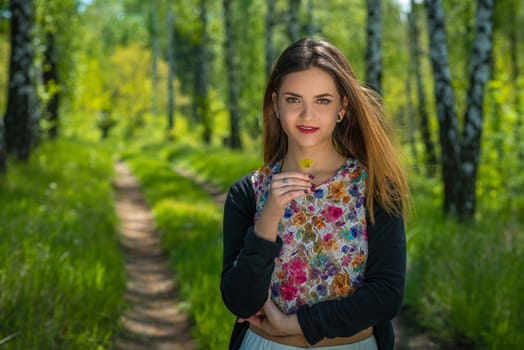 Young woman with red lips and brown hair is holding one yellow dandelion in hand while standing on a footpath in a forest during spring time.