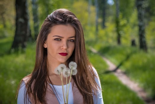 Young woman with red lips and brown hair is holding five white dandelions in hand while standing on a footpath in a forest during spring time.