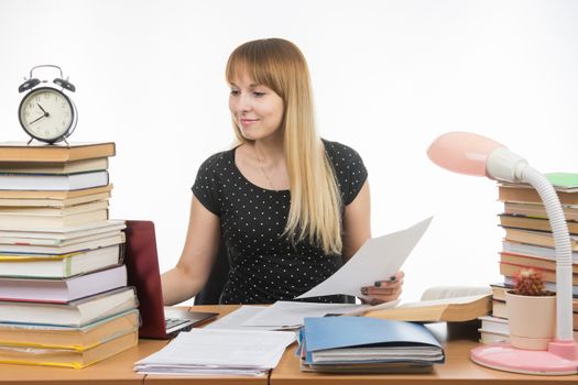 Joyful girl behind a desk littered with books, papers in hand looking in laptop