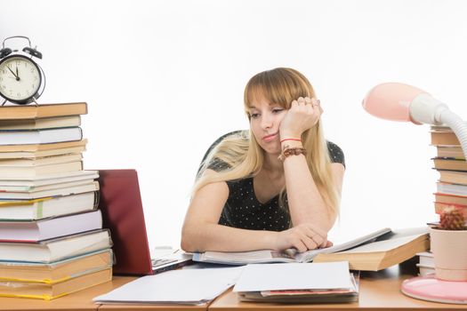 Student sadly looks at the laptop at the table among the stacks of books and papers