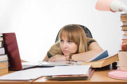 The girl at the desk crammed with books and papers sadly looks at the laptop with her head resting on his hand on the table
