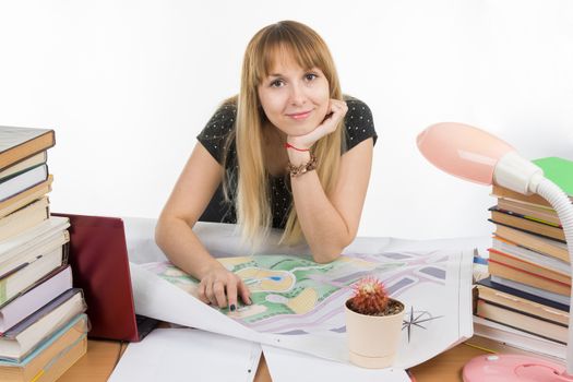 A girl student at a desk littered with books and drawings with a smile looks in the frame