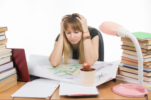A girl student at a table with a pile of books, drawings and projects sits holding his head in his hands