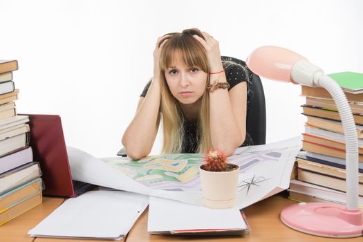 Student designer behind a desk with a pile of books and projects sits holding his head in his hands
