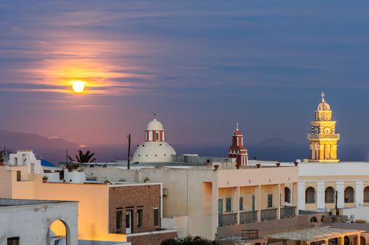 Illuminated buildings, domes and towers of of Fira at night, Santorini, Greece. Moon at sky.