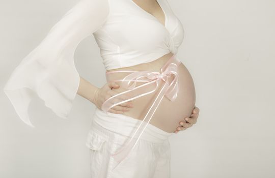 Torso of a pregnannt woman with pink ribbin on her abdomen dressed in white on gray background.