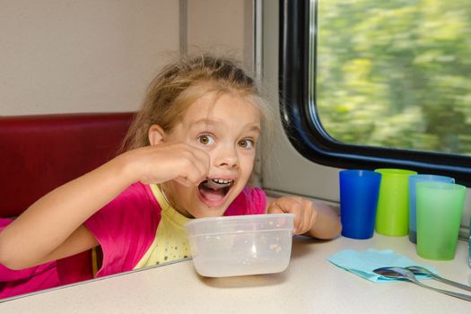 The girl on the train sits at a table on the lower place in the second-class compartment of the car and eats it with relish spoon