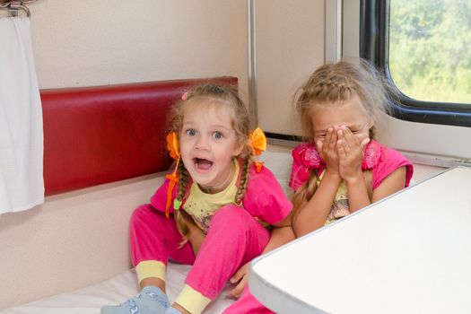 Two cheerful little girls are sitting on the train on the lower second-class place car in the same pajamas