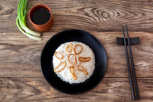 Spicy stir fried squid with rice and green onion and chopstick on black bowl on rustic brown wooden background