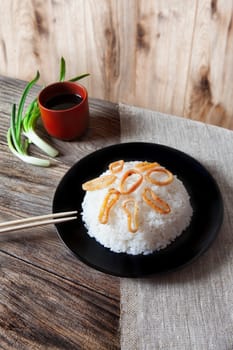 Spicy stir fried squid with rice and green onion and chopstick on black bowl on rustic brown wooden background