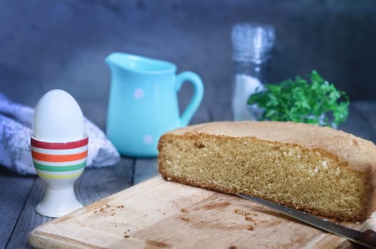 Half of bread on a cutting Board, knife and egg in a striped stand. In the background: blue pitcher, salt shaker,napkin and bunch of greens. Rustic style, focus, bokeh.