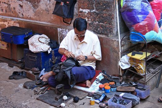 A man repairs rucksack on the footpath against building.Cobbler tools and necessities are around him. Vasco da Gama town, Goa, India.