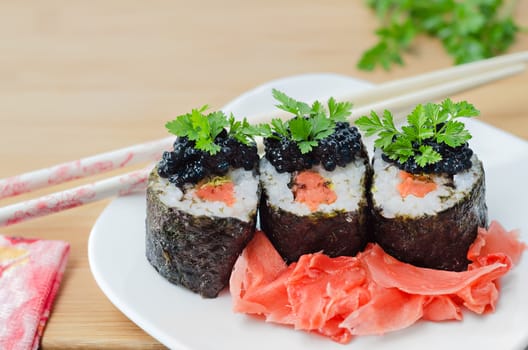 Rolls with salmon, caviar and pickled ginger, decorated with greenery on white plate and wooden background.