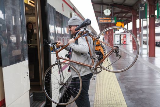 A young man with glasses and a hat is coming into a wagon-train with his bike in a urban scene