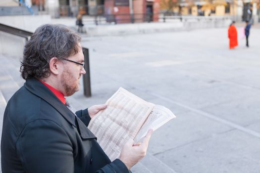 A young businessman with glasses reading a newspaper is thinking and talking by smartphone  in urban scene