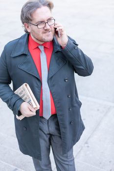 A young businessman with glasses reading a newspaper is thinking and talking by smartphone  in urban scene