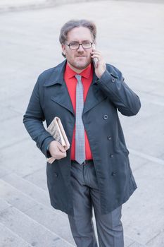 A young businessman with glasses reading a newspaper is thinking and talking by smartphone  in urban scene