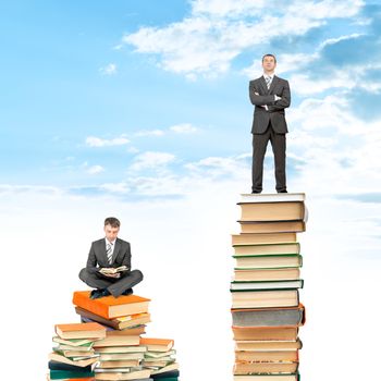 Businessman sitting on stack of books and reading opened book, another man standing on books