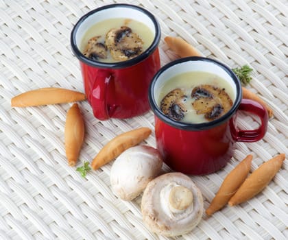 Arrangement of Delicious Homemade Mushrooms Cream Soup Decorated with Roasted Champignons in Red Cups and Bread Sticks closeup on Wicker background
