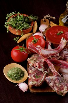 Arrangement of Raw Lamb Ribs with Tomatoes, Garlic, Herbs, Spices and Olive Oil on Wooden Cutting Board closeup on Dark Wooden background