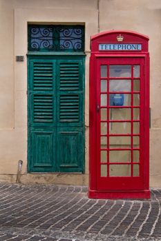 Traditional english style of a red telephone cabin. In the background green old gate. Street of fishing village Marsaxlokk on the island Malta.