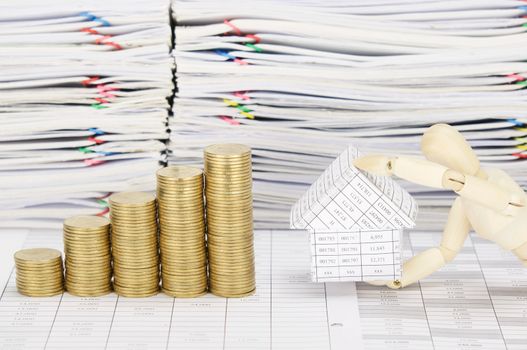 Wooden dummy holding house with step pile of gold coins on finance account have blur overload of paperwork with colorful paperclip as background.