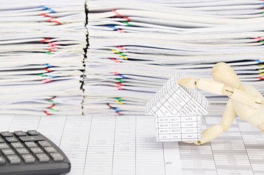 Wooden dummy holding house on finance account have blur calculator and overload of paperwork with colorful paperclip as background.