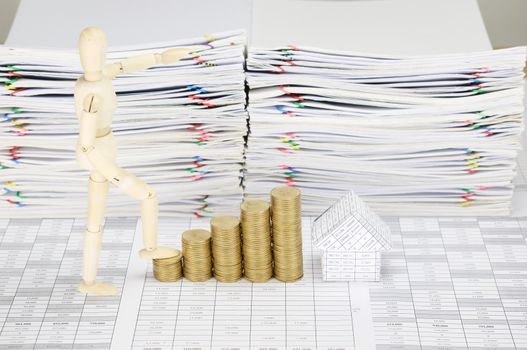 Wooden dummy step up with step pile of gold coins and house on finance account have blur overload of paperwork with colorful paperclip as background.