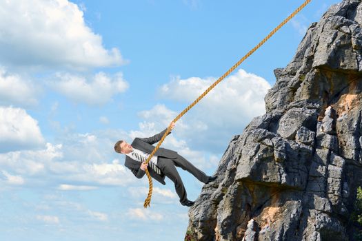 Young businessman scaling rock with rope, cloudscape