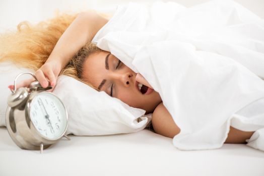 Young woman lying in bed in the morning. She is waking up with alarm clock.