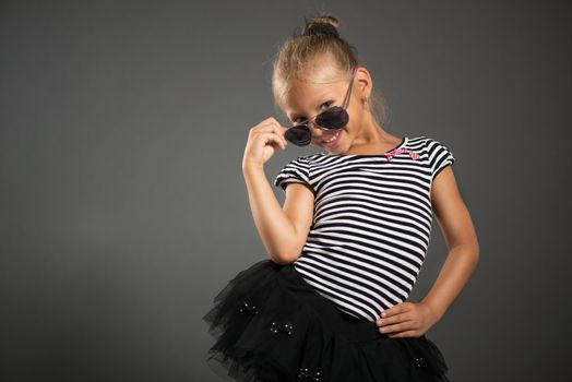 Pretty little girl smiling and posing with sunglasses. Studio shutting. Grey background. Looking at camera.