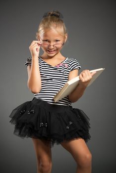 Pretty little girl smiling and posing with book. Studio shutting. Grey background. Looking at camera.