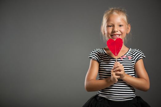 Pretty little girl smiling and posing with lollipop. Studio shutting. Grey background. Looking at camera.