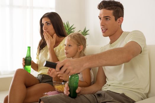 Beautiful Happy young mother and father and their cute daughter sitting at home and watching movie.