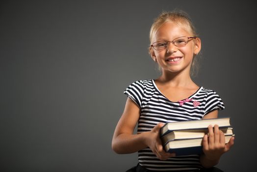 Cute little girl standing and holding books. Studio shutting. Grey background. Looking at camera.
